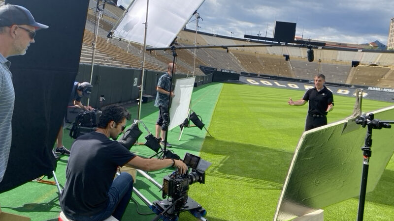 Snowy Peak Films crew filming a Wilhite Law Firm commercial on the field at Folsom Field in Boulder, with a cinema camera on a dolly, diffusion, bounce boards and a boom mic