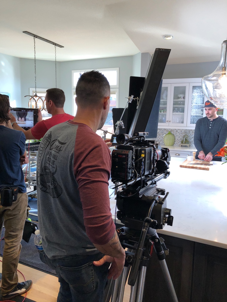 Cinema camera on a tripod framing talent at a kitchen island while the crew checks the shot on a monitor, filming a commercial for Nature Raised Farms